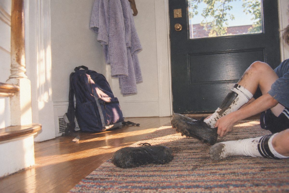 A boy pulling off muddy soccer cleats in a sunny hallway