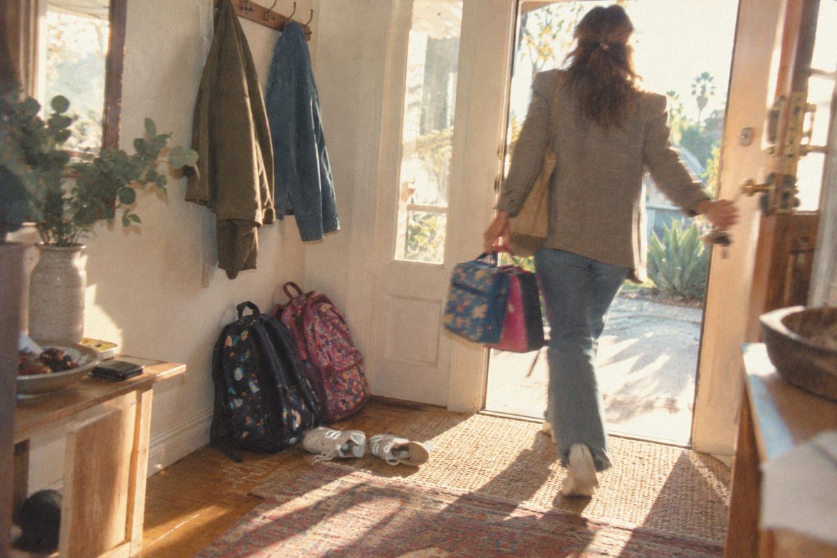 A woman heading out the front door with two lunch boxes, backpacks left behind